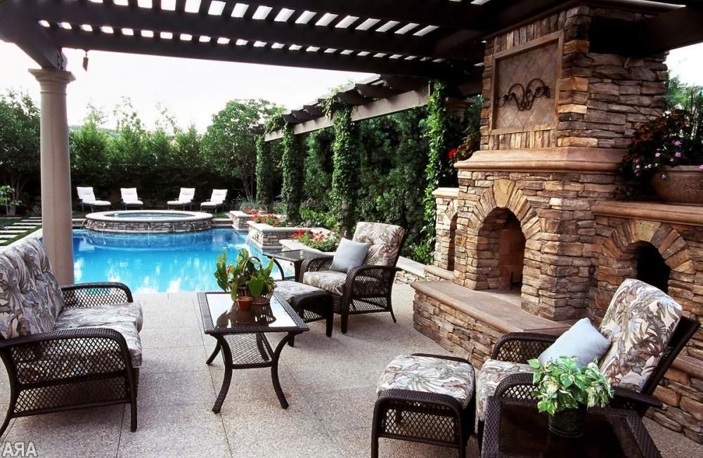 Patio lounge with wicker chairs, stone fireplace, and a pool under a pergola