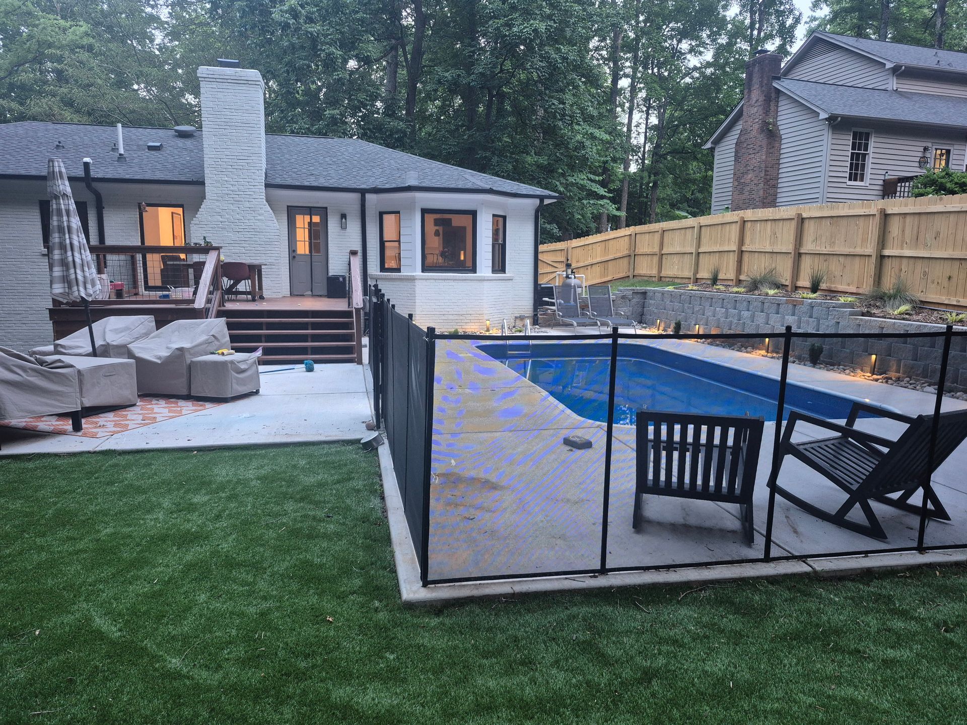 Backyard patio with a lit house, stone fireplace, pool, deck chairs, and fenced landscaping at dusk