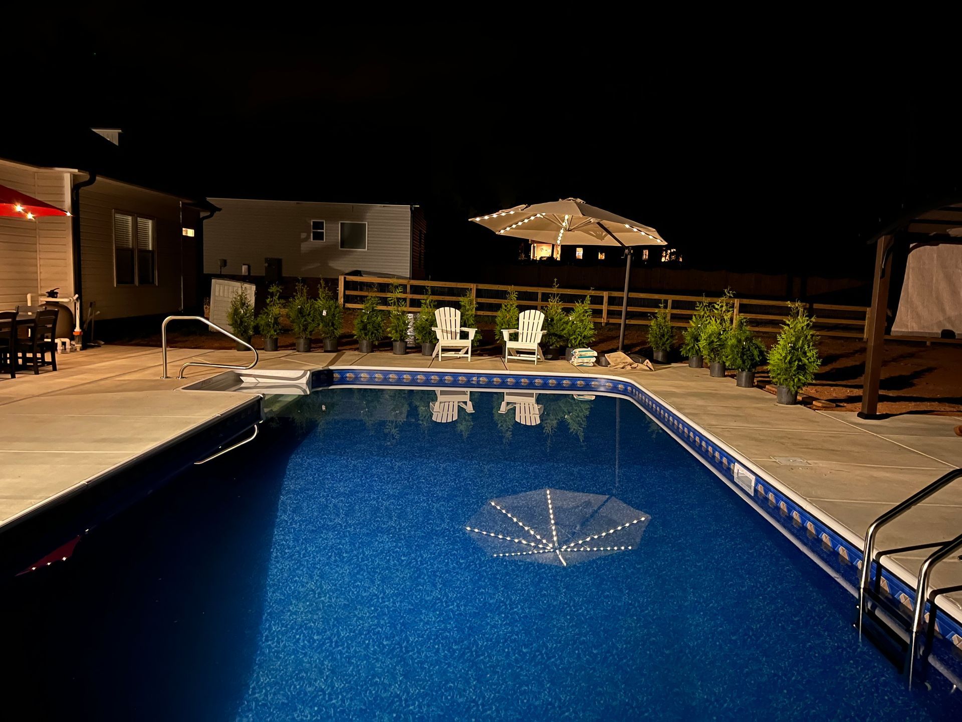 Nighttime backyard pool with blue water, patio lights, lounge chairs, and a lit umbrella.