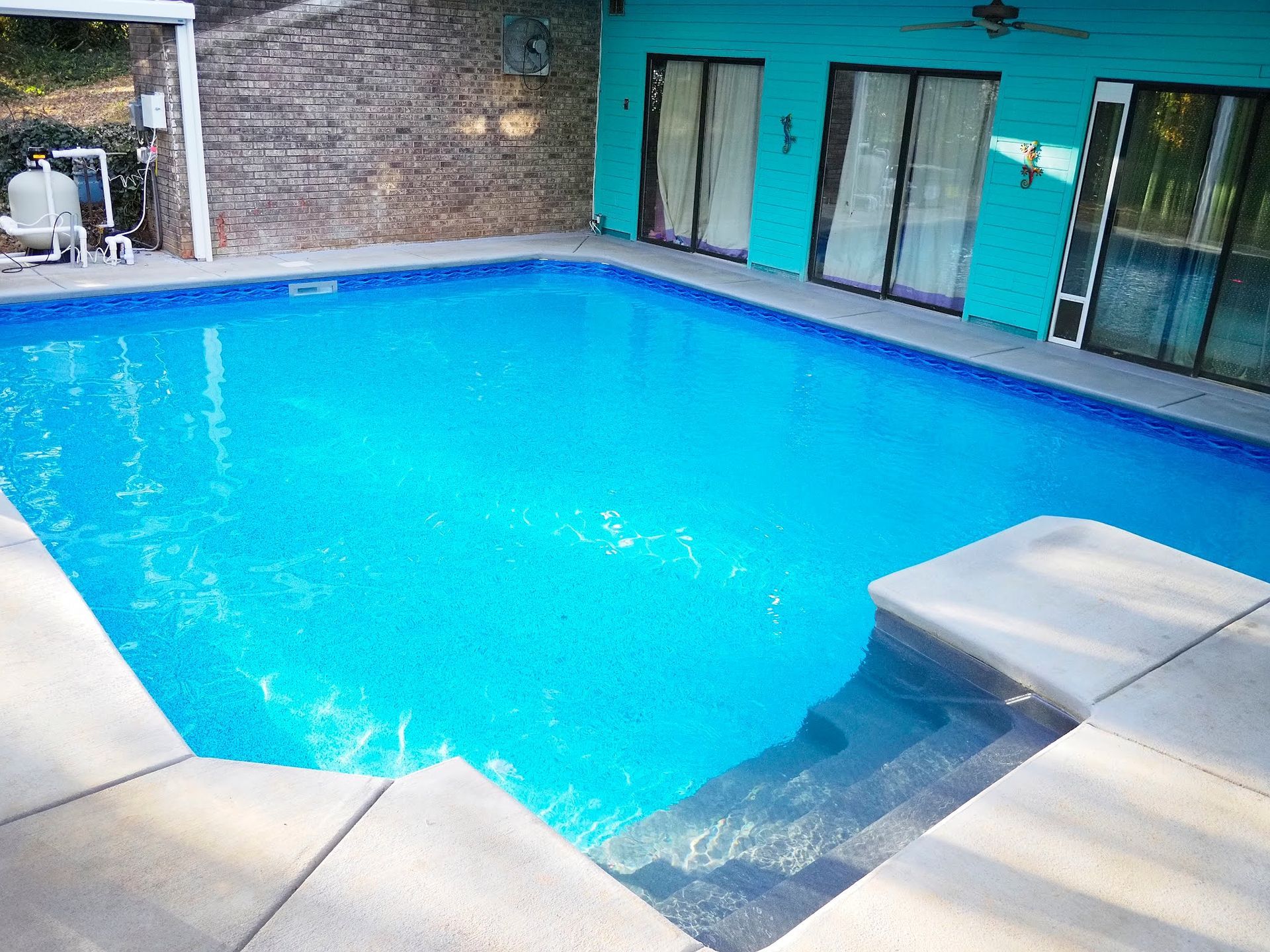 Bright blue swimming pool with a small shallow ledge beside a brick patio and turquoise wall