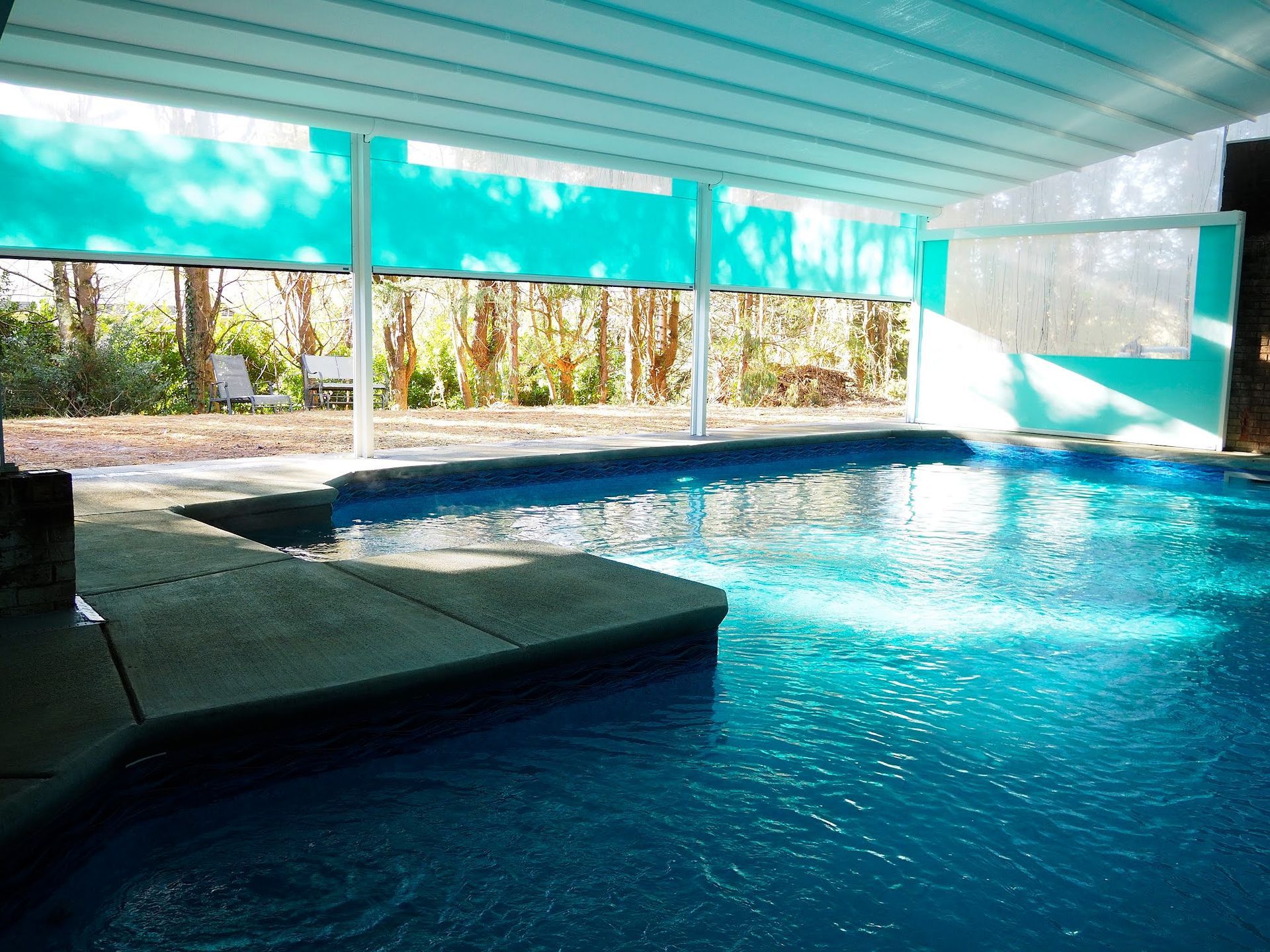 Covered indoor pool with blue water and a concrete diving platform, lit by bright natural light.