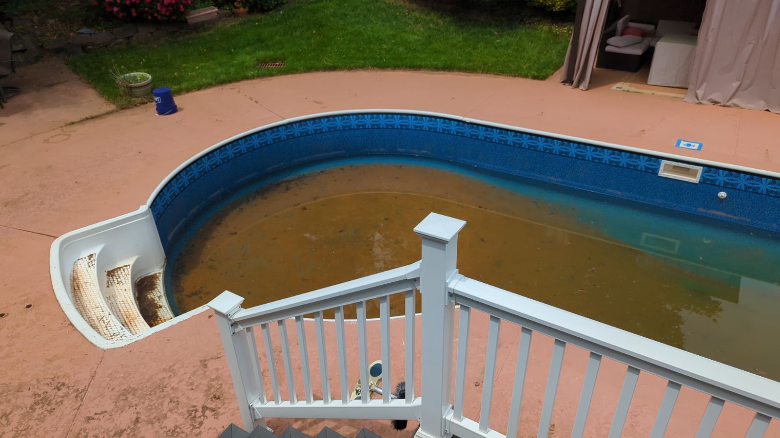 Partially drained backyard pool with brown water, blue lining, and a white fence in the foreground