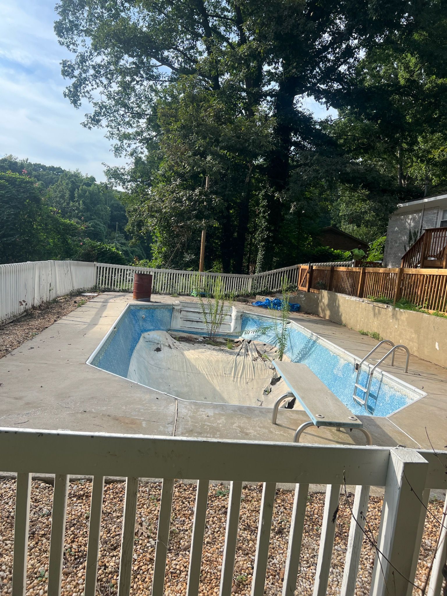 Empty abandoned blue-and-white pool enclosed by a fence, with trees in the background