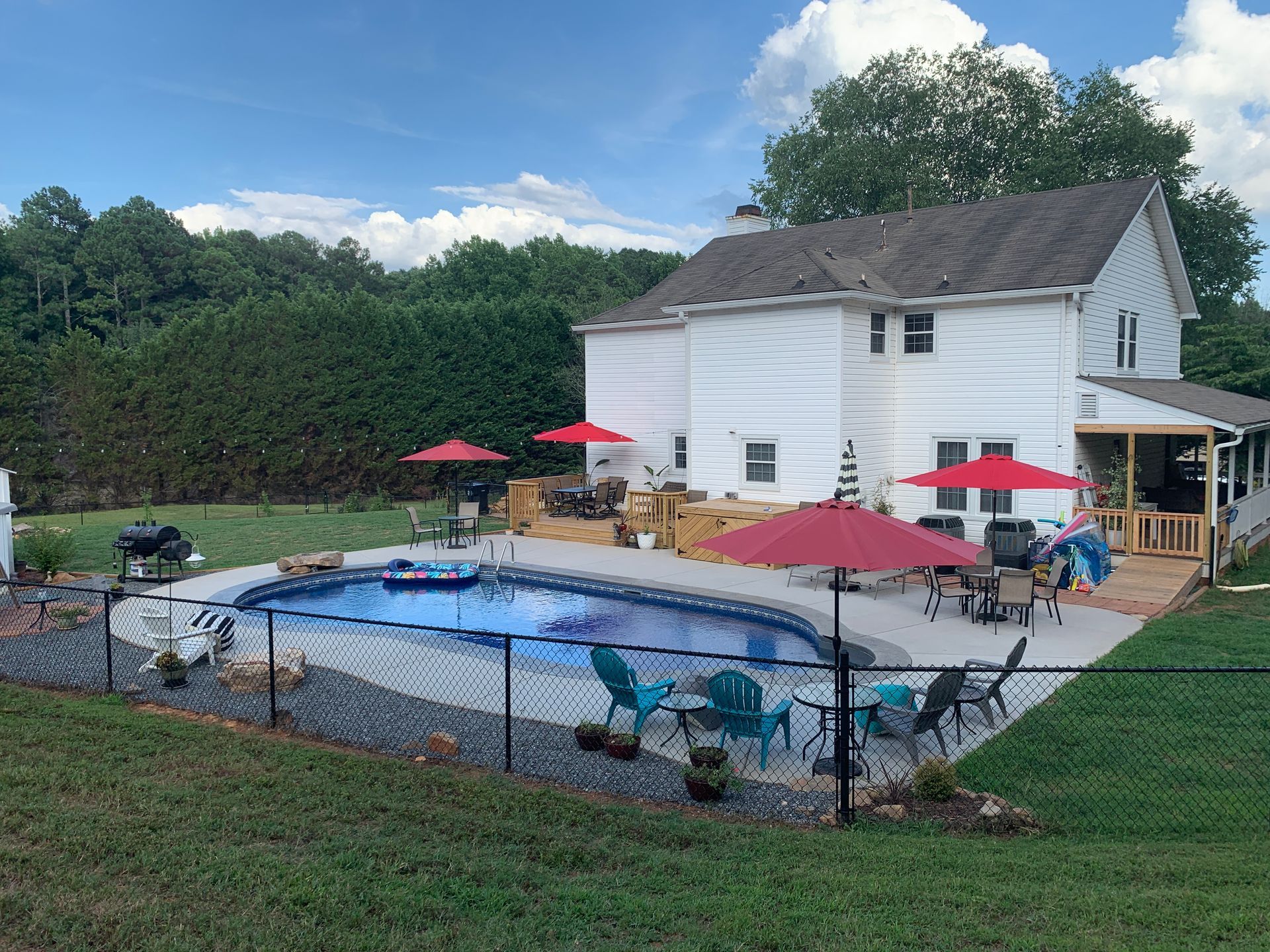 Backyard pool beside a white house, with red umbrellas, deck chairs, and a fenced lawn.