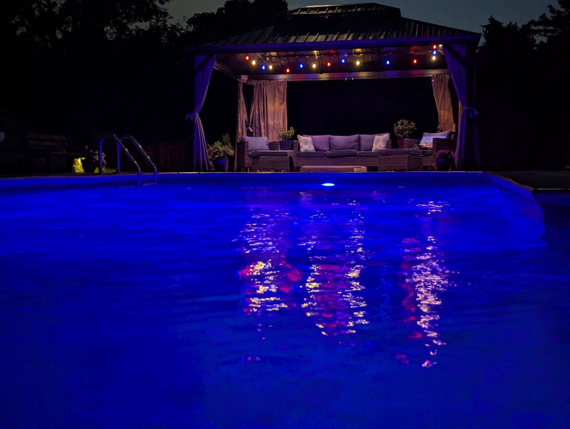 Blue-lit backyard pool at night with a lit cabana and reflections on the water