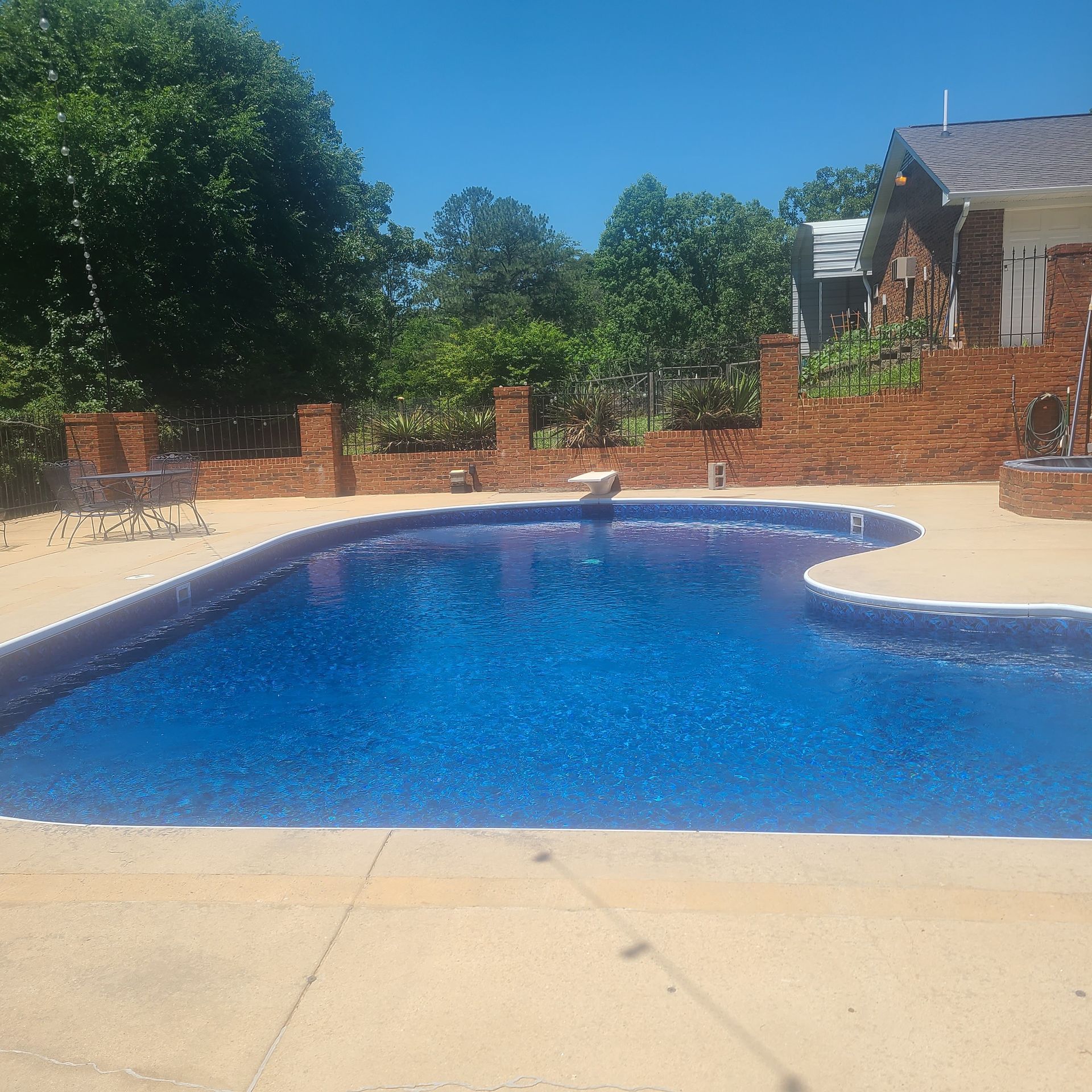 Backyard swimming pool with blue water, brick patio, and trees under a clear sky