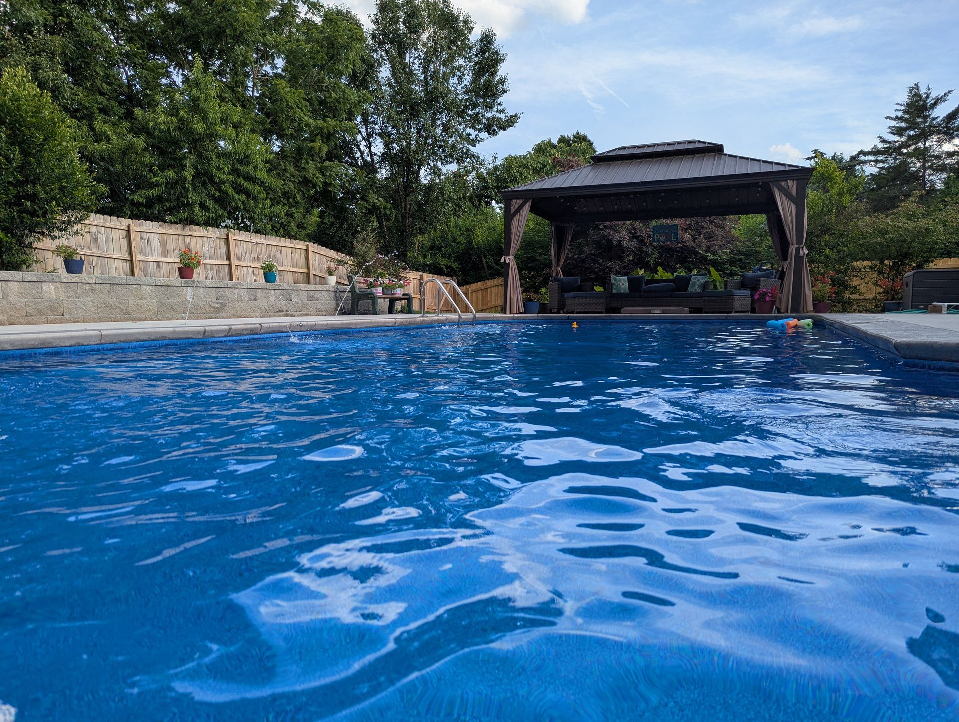 Blue swimming pool with a stone wall and shaded gazebo under a partly cloudy sky