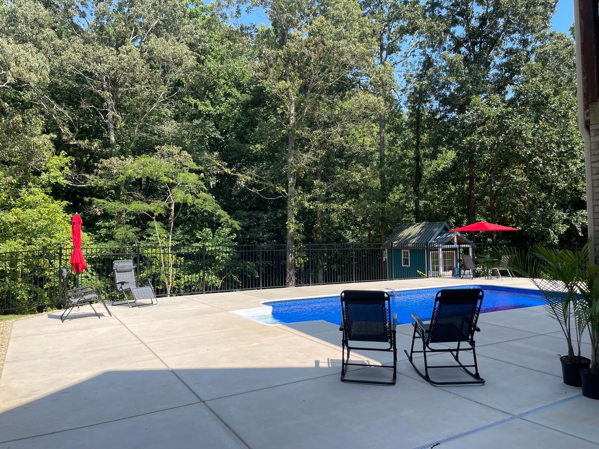 Backyard pool with blue water, patio chairs, red umbrellas, and trees in the background