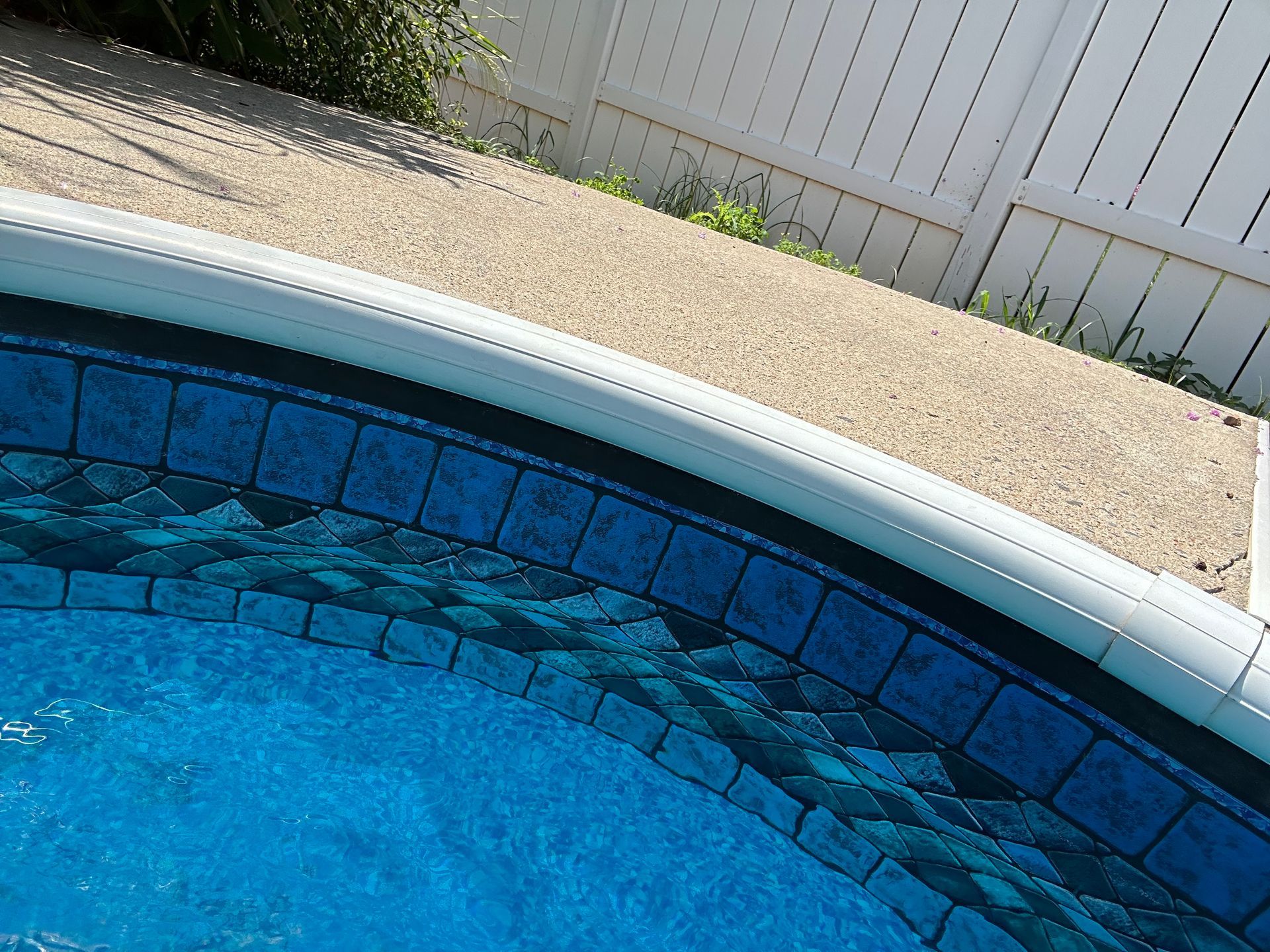 Curved blue swimming pool edge beside a gravel deck and white fence
