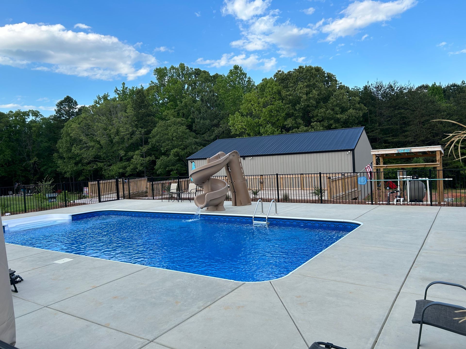 Swimming pool with blue water, tan deck, and a small slide beside a building and trees.
