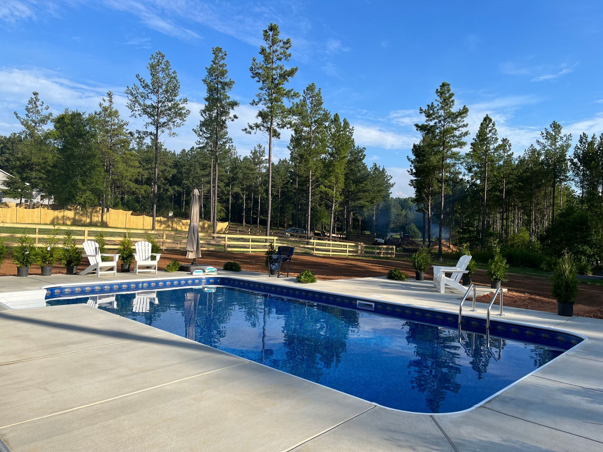 Backyard pool with blue water, stone deck, white chairs, and pine trees under a clear sky