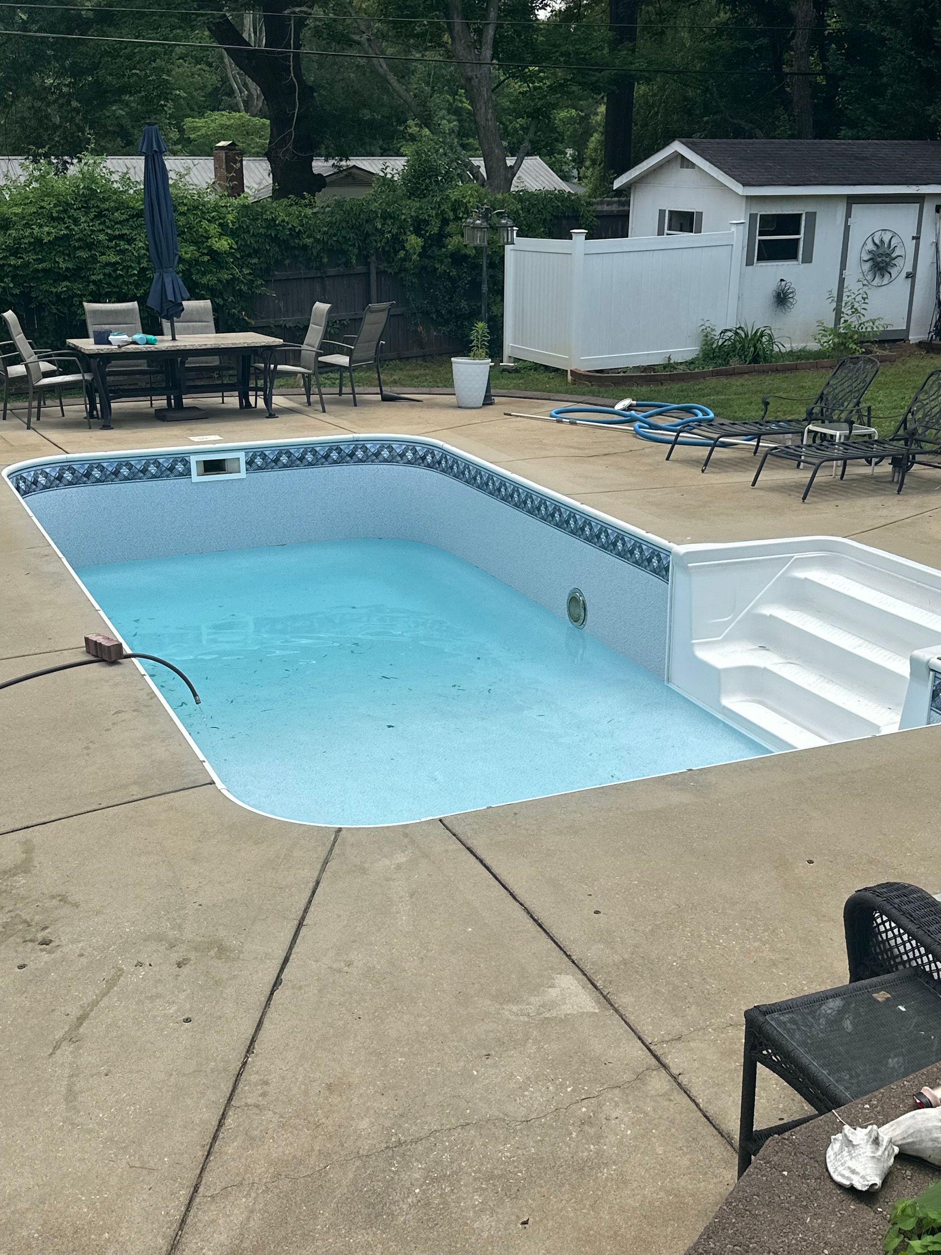 Backyard pool with white steps, tan deck, lounge chairs, and a table under trees nearby.