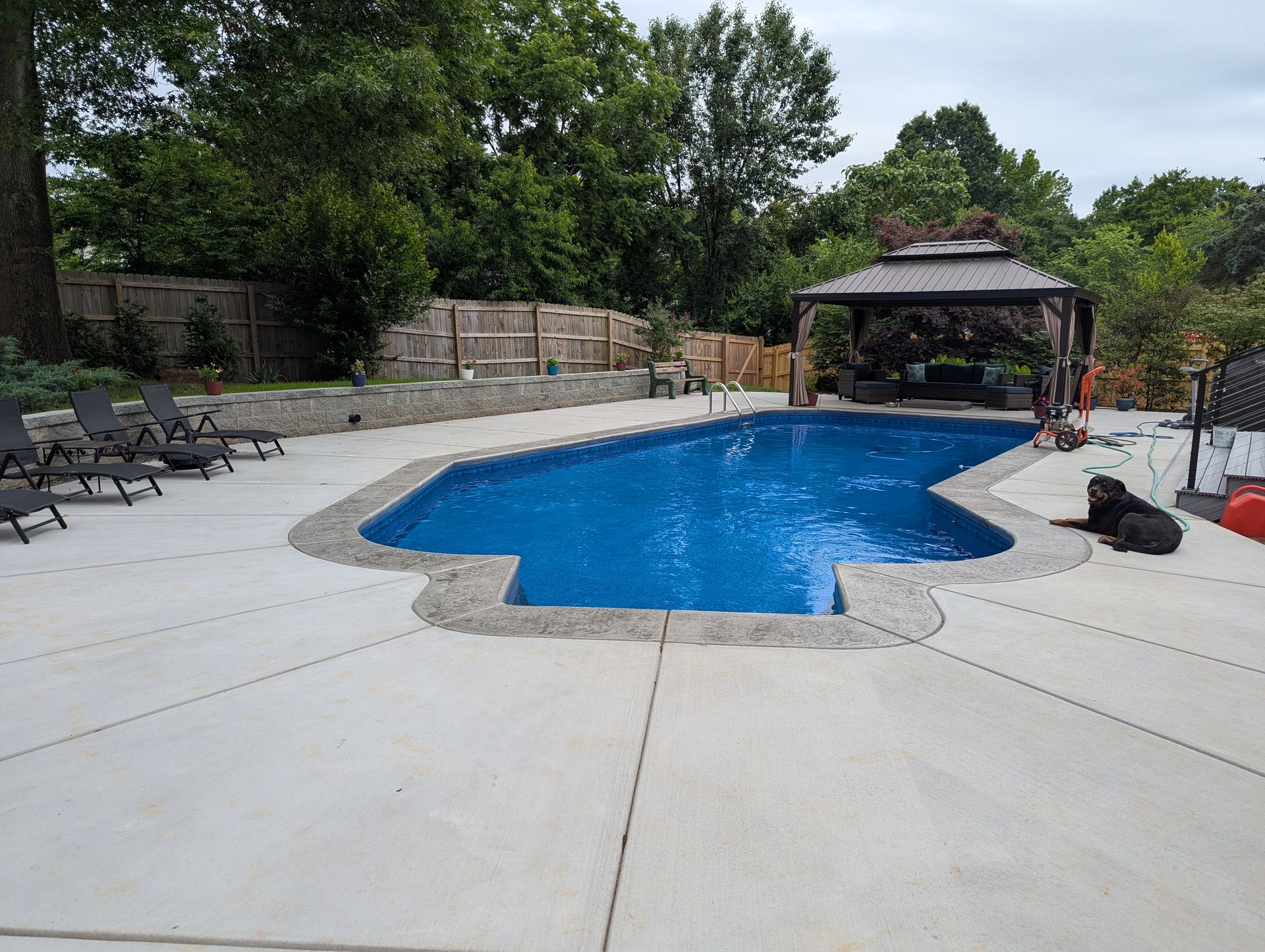 Backyard swimming pool with blue water, patio chairs, stone wall, and gazebo amid trees