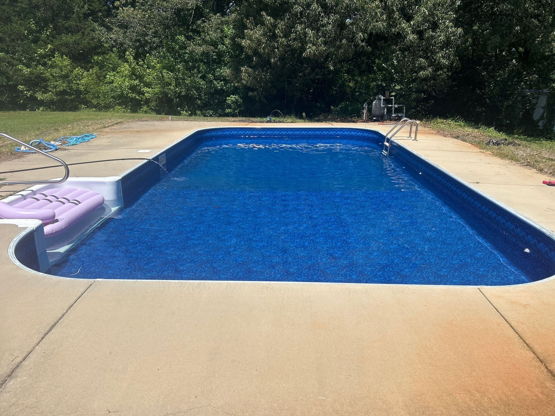 Backyard swimming pool with blue water and tan concrete deck, surrounded by trees