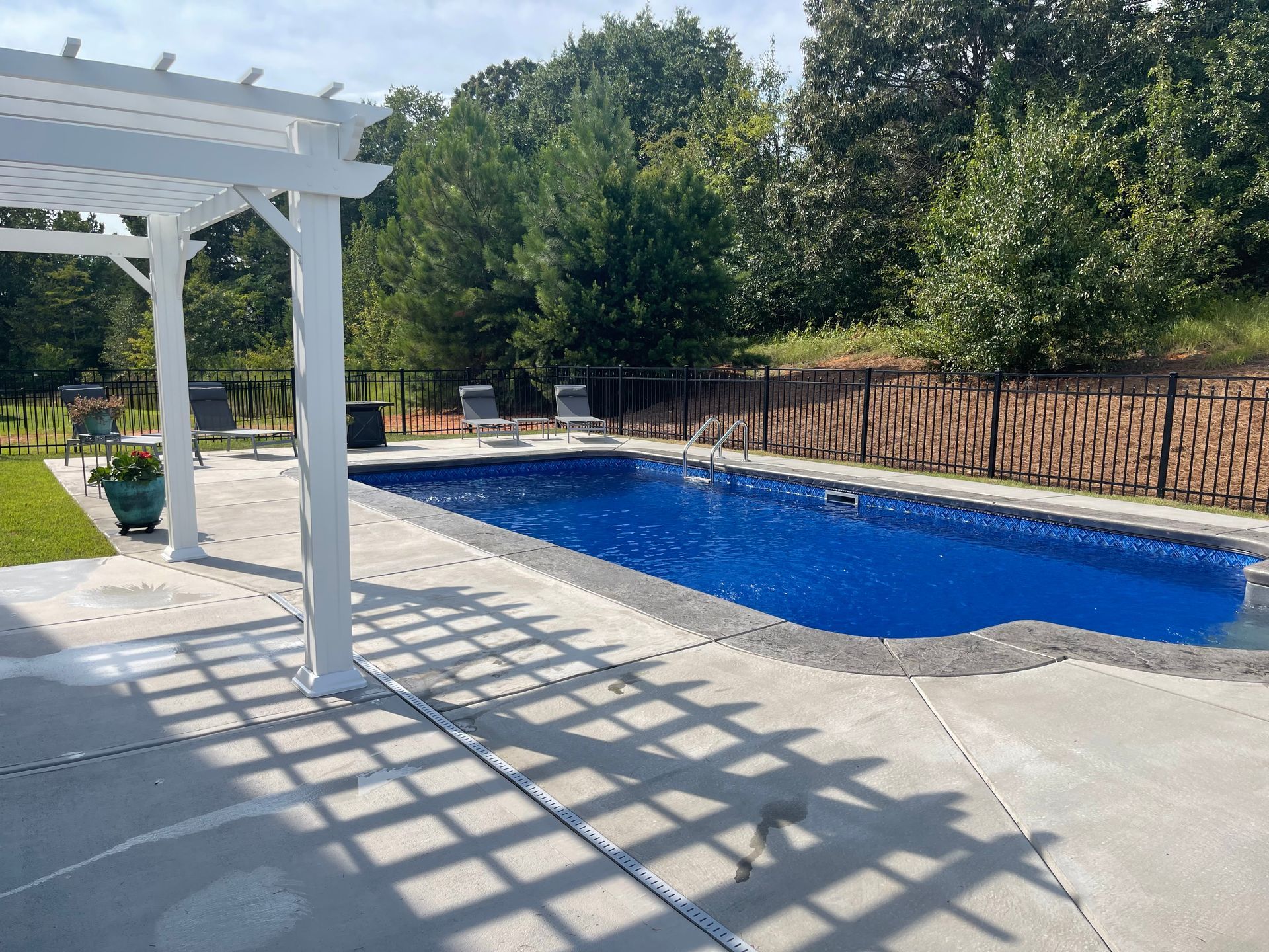 Backyard pool beside a white pergola and deck, with trees and a wooden fence in the background.