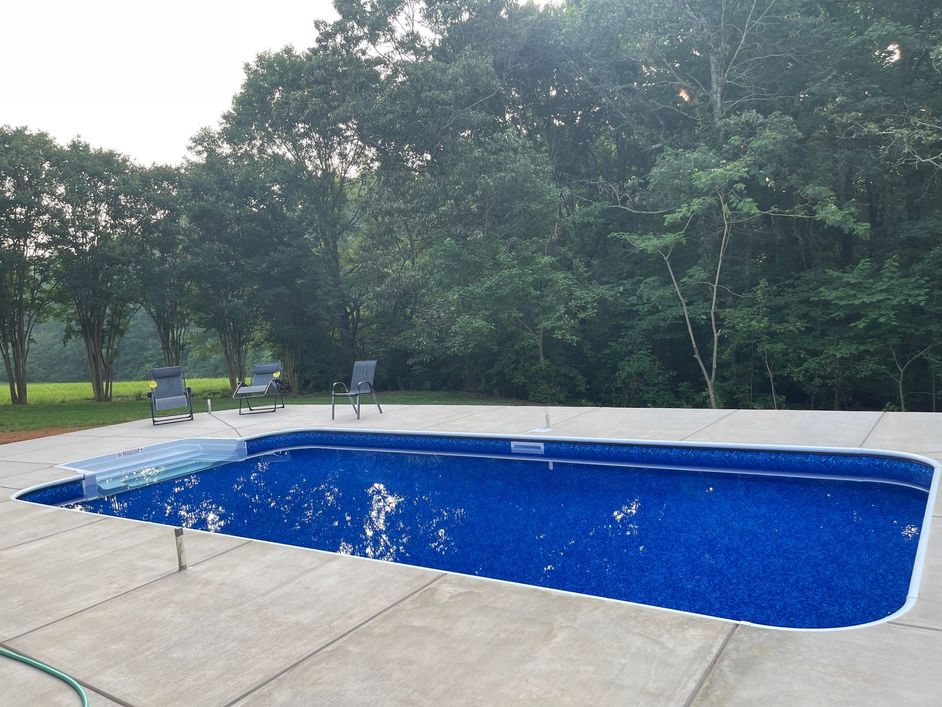 Rectangular blue in-ground pool with water jets, bordered by concrete patio and trees