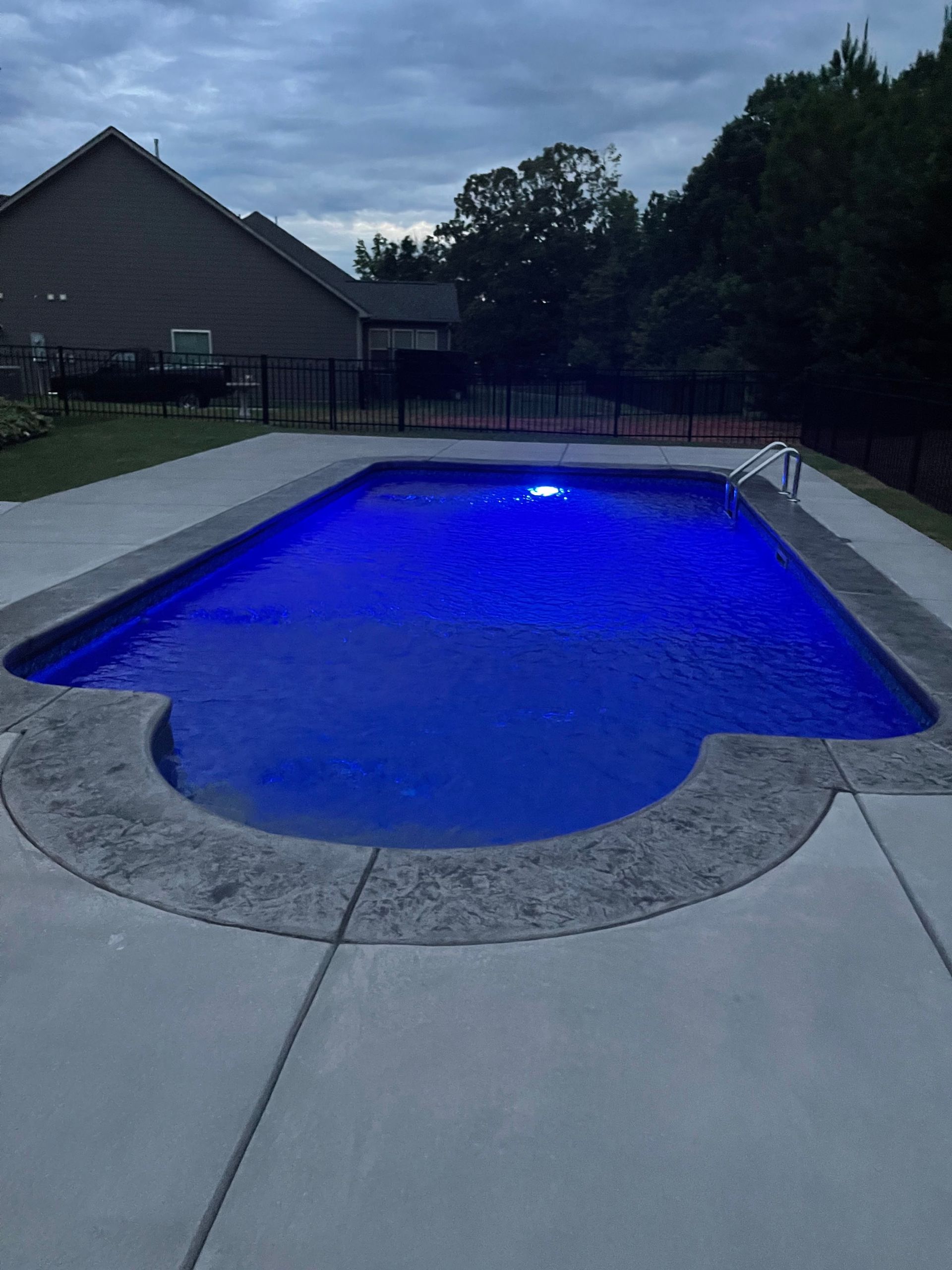 Backyard swimming pool glowing blue at dusk, with concrete deck and houses in the background