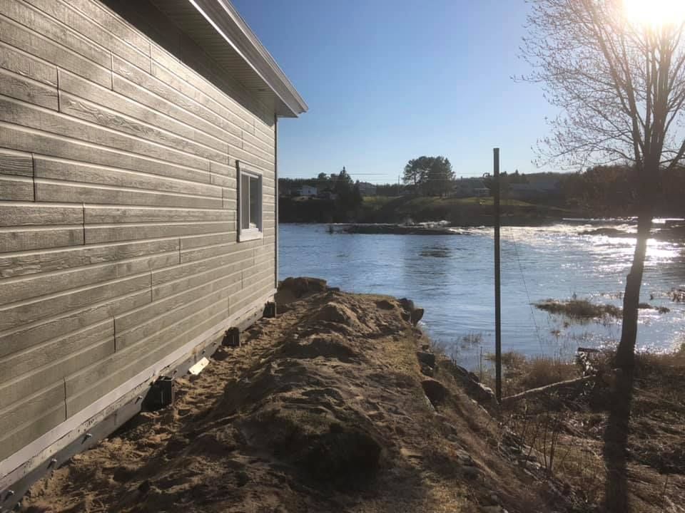 Vue latérale d'une maison claire longeant une rive et l'eau sous un soleil éclatant.