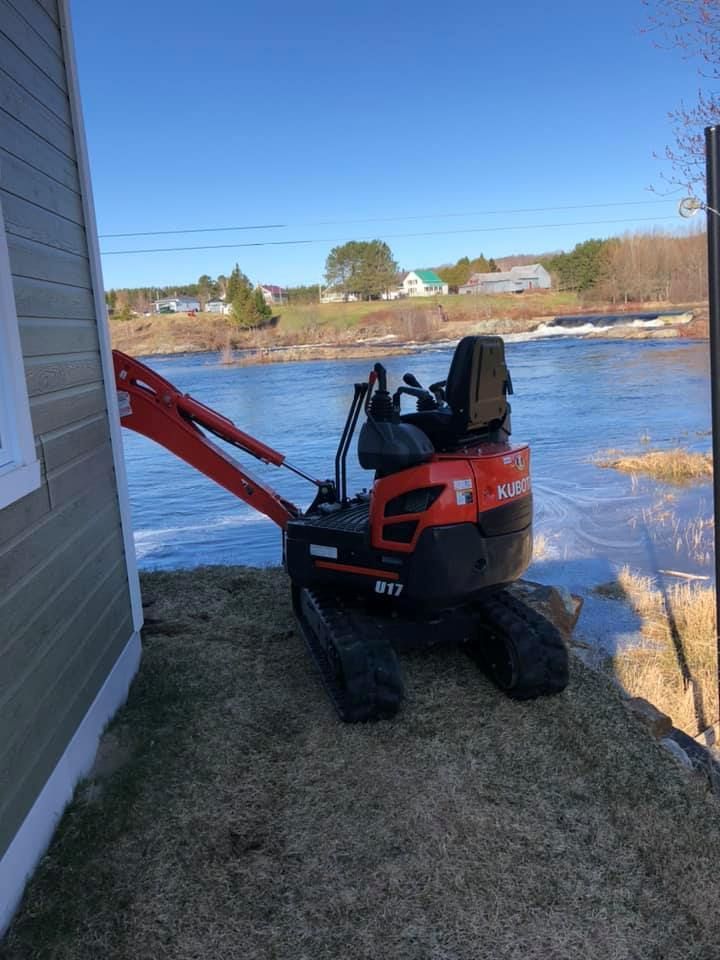 Une mini-pelle Kubota U17 orange est garée sur un terrain herbeux à côté d'une maison, avec vue sur une rivière par une journée ensoleillée.