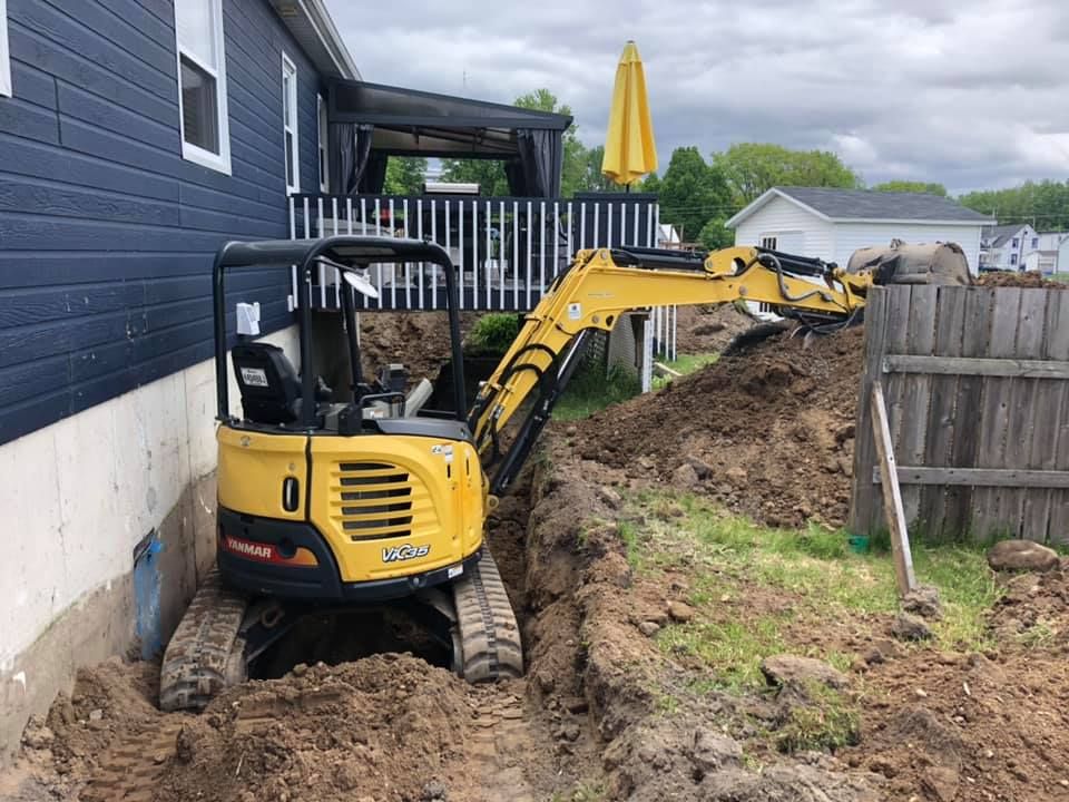 Une mini-pelle jaune creuse une tranchée le long des fondations d'une maison bleue, dans un jardin à côté d'une clôture en bois.