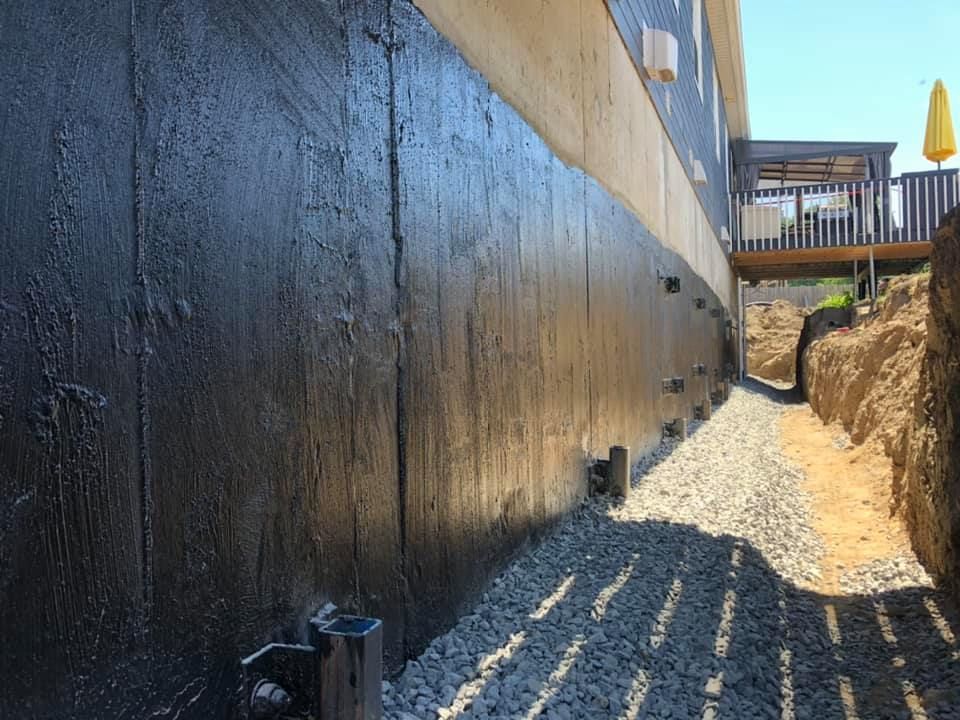 Un mur de fondation de maison avec un revêtement imperméable noir, une base de gravier et une tranchée excavée à côté.