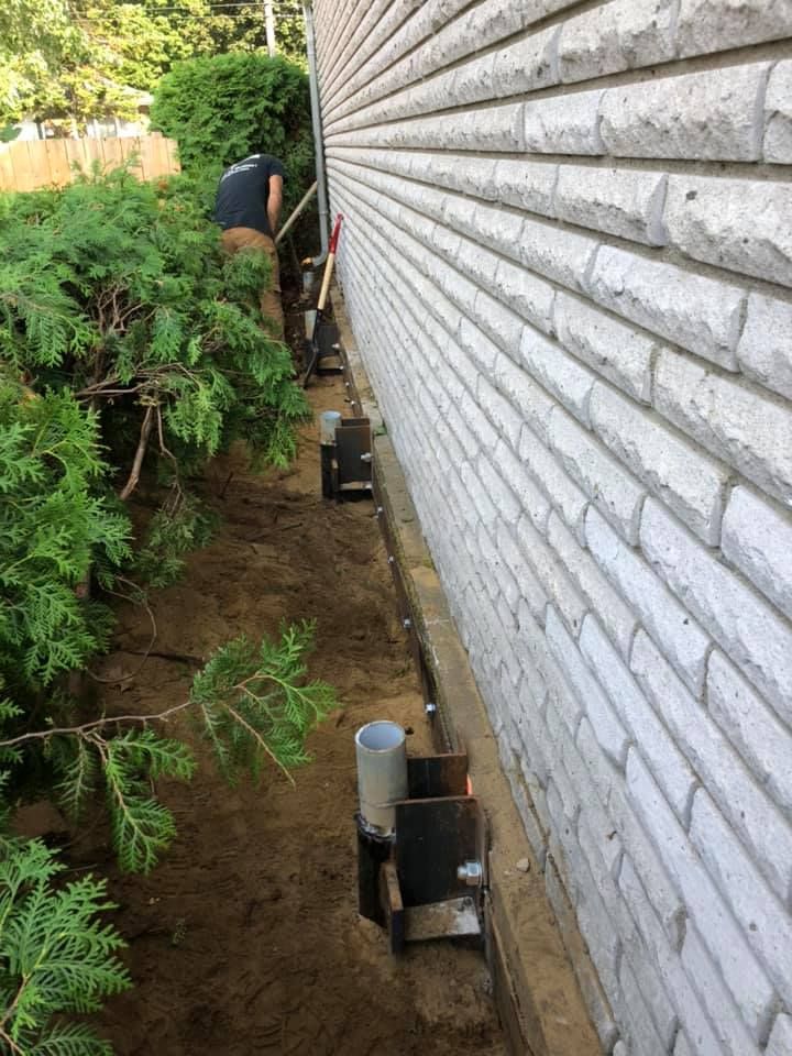 Une personne creuse le sol à côté des fondations d'une maison, où plusieurs supports structurels en acier sont boulonnés au mur.