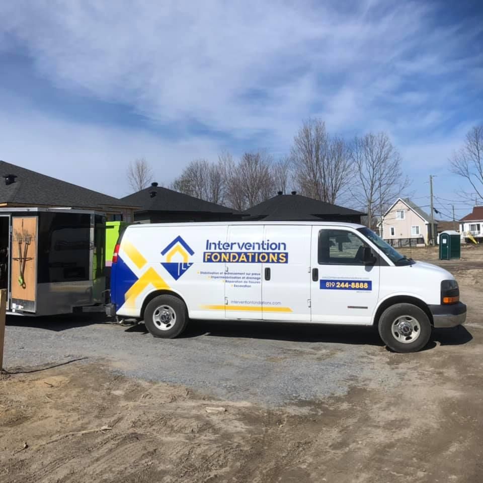 Une camionnette de service blanche portant le logo « Intervention Fondations » tire une remorque sur un terrain gravillonné sous un ciel bleu azur.