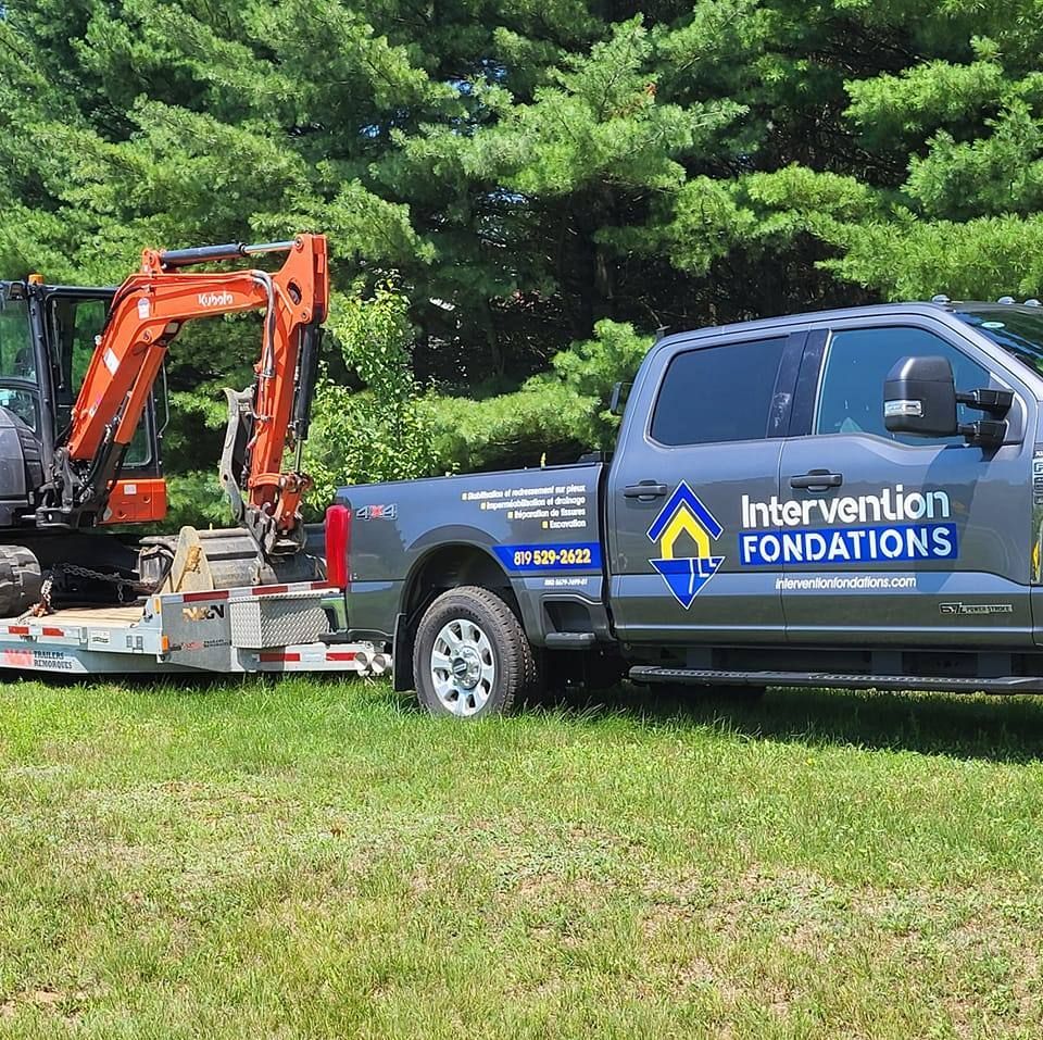 Une camionnette grise des Fondations Intervention tractant une excavatrice de chantier orange sur une remorque près d'arbres.