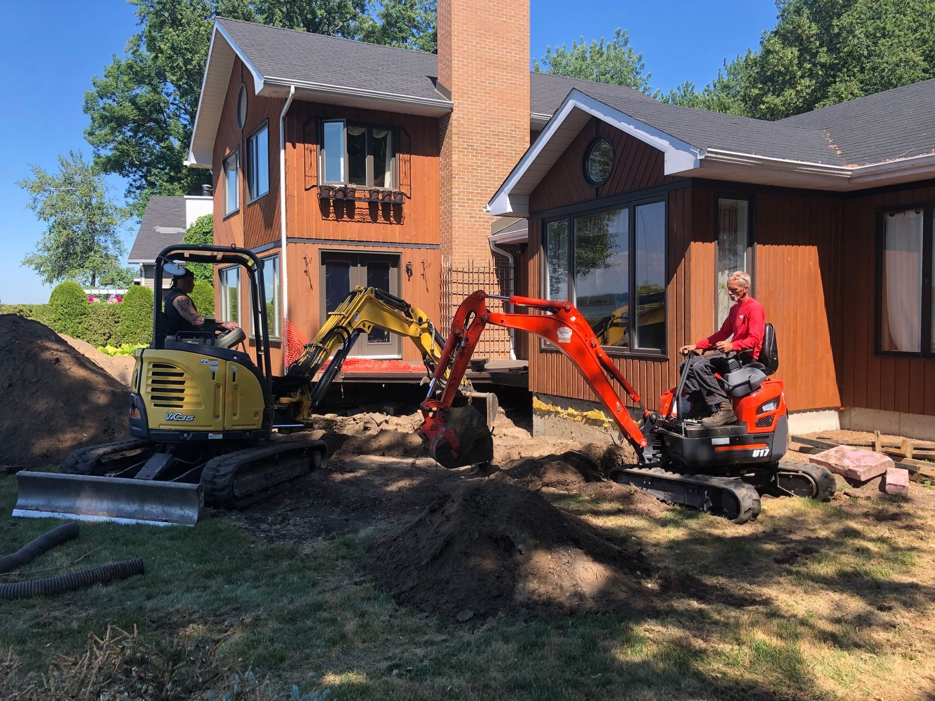 Deux ouvriers du bâtiment manœuvrent des mini-pelles jaunes et orange devant une maison en bois brun à plusieurs étages.