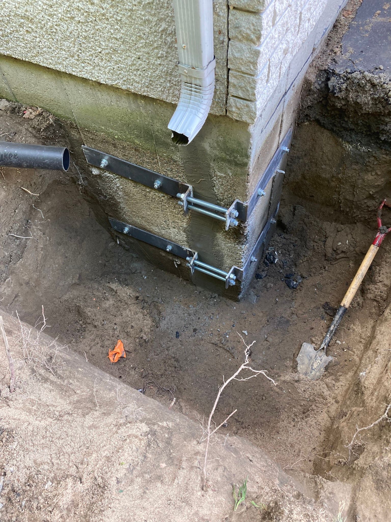 Un coin des fondations d'une maison, avec des supports en acier installés dans une zone de terre excavée, et une pelle à proximité.