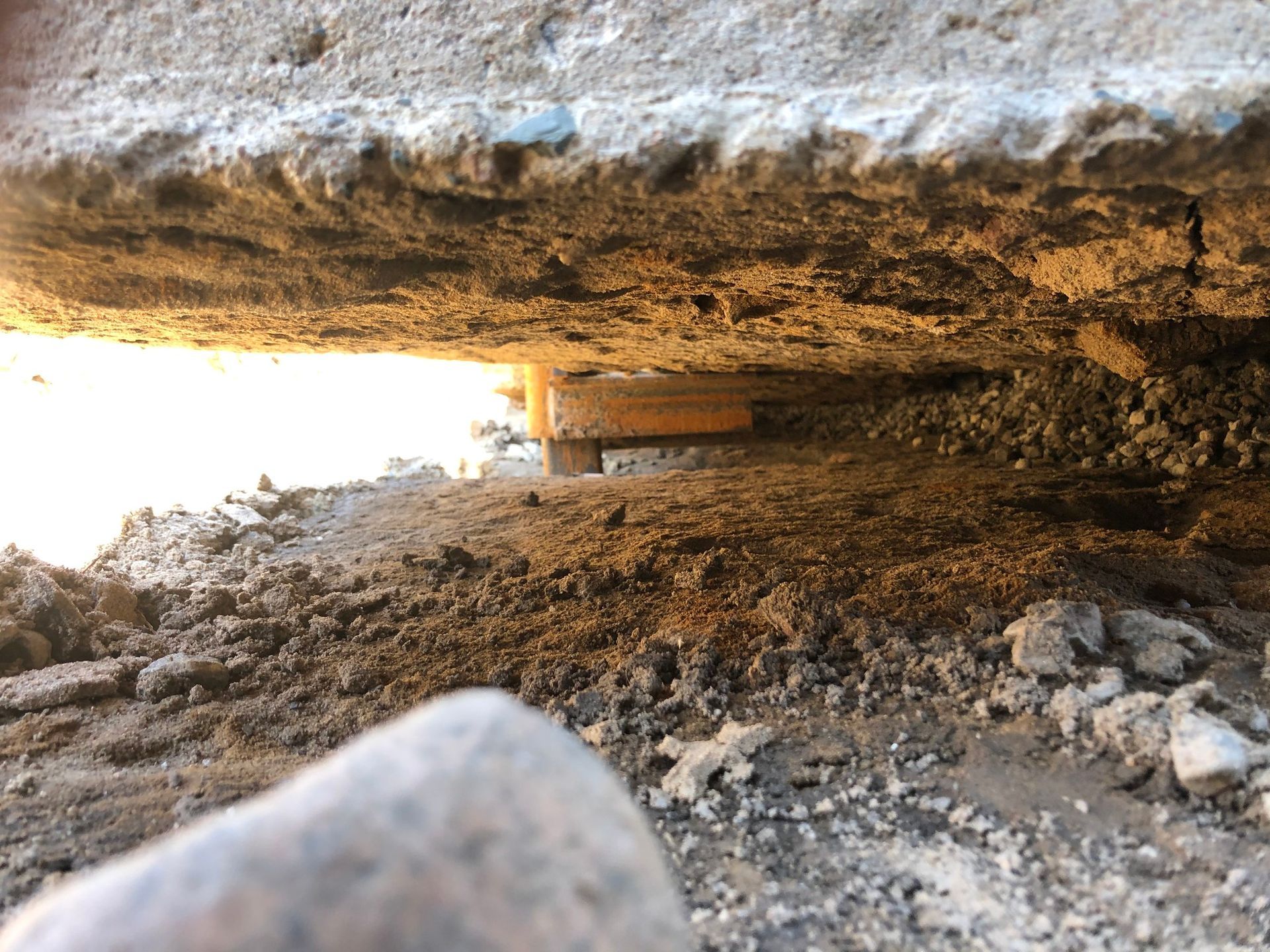Vue au niveau du sol sous une grande dalle de béton, révélant la terre, les rochers et un petit poteau de soutien en bois.