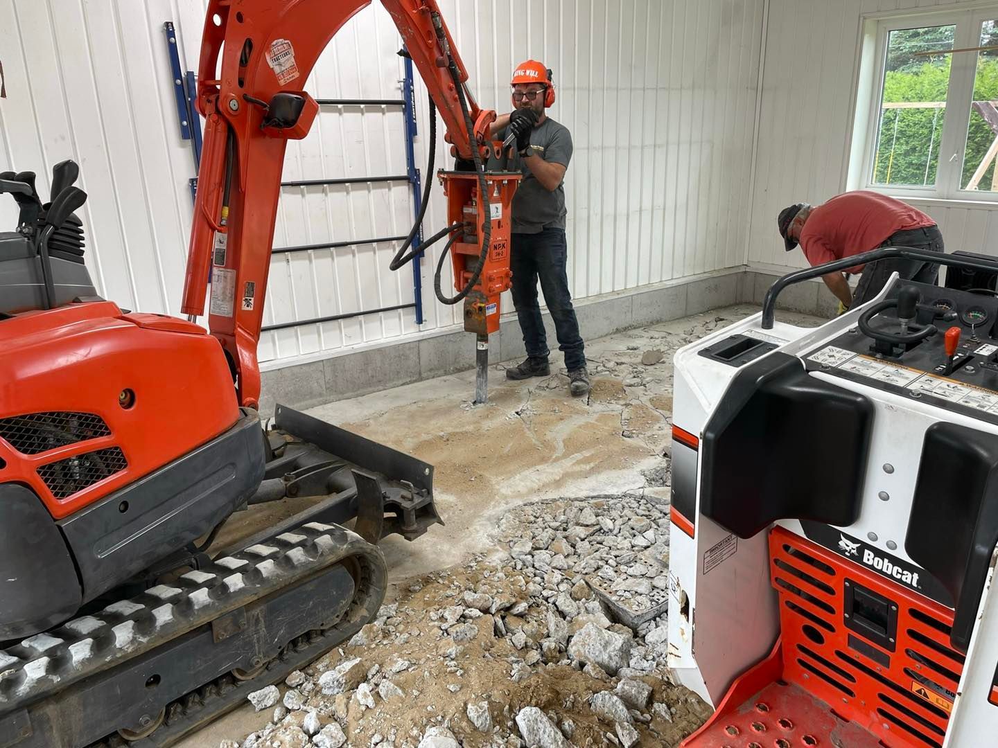 Un ouvrier utilise une excavatrice orange équipée d'un marteau hydraulique pour démolir un sol en béton à l'intérieur d'une pièce lumineuse.