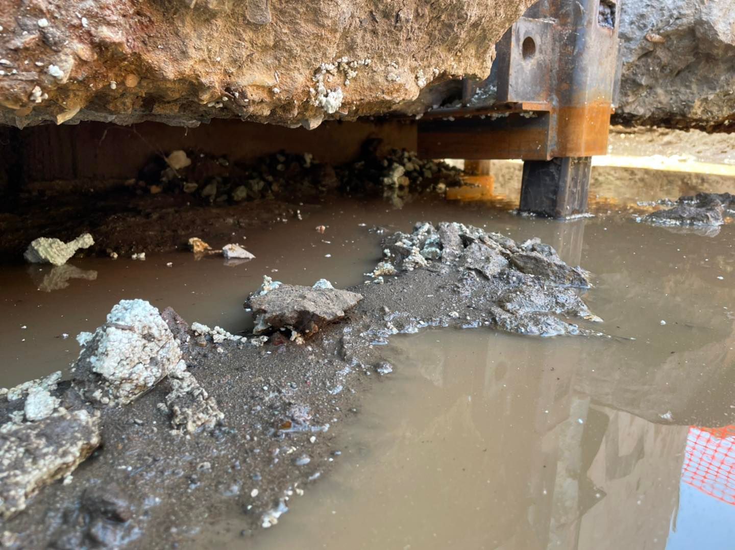Un chantier de construction montrant une dalle de fondation en béton suspendue au-dessus d'une tranchée remplie d'eau boueuse, avec un poteau en acier rouillé.