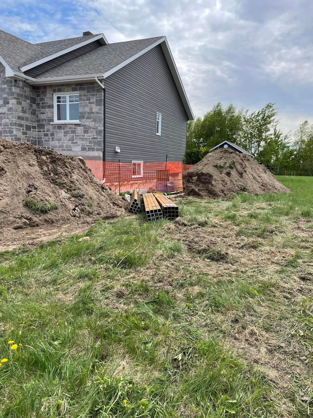 Une maison en construction, avec un revêtement en pierre et gris, des tas de terre excavée et des piles de planches de bois près du mur.