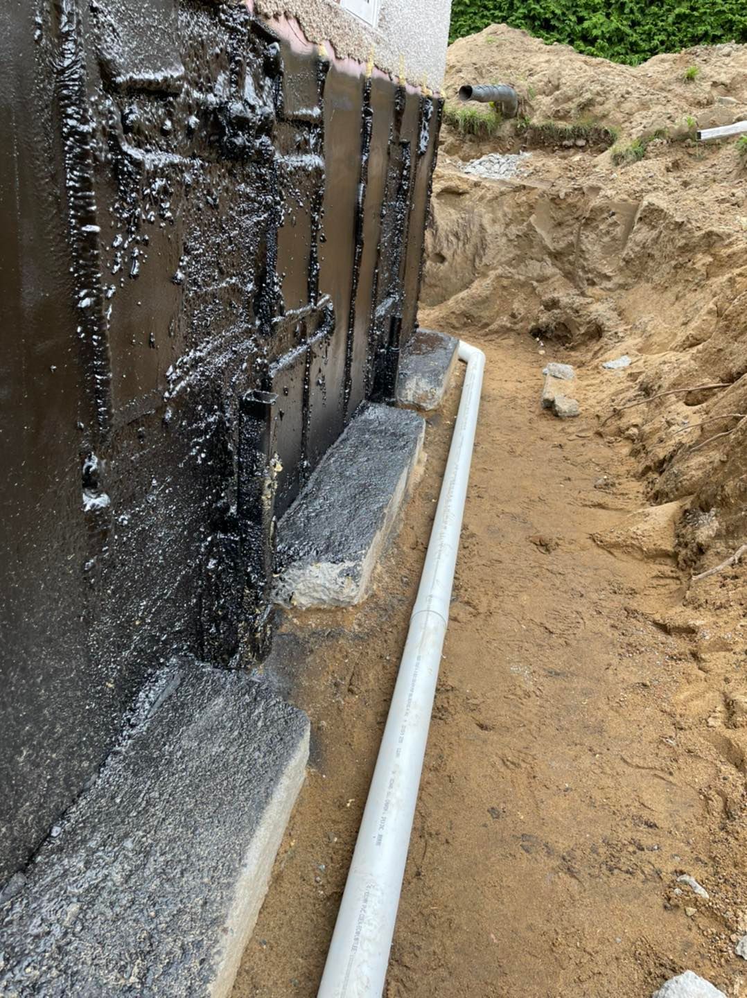 Un mur de fondation recouvert d'un revêtement imperméable noir se trouve à côté d'un tuyau de drainage blanc dans une tranchée de construction sablonneuse.