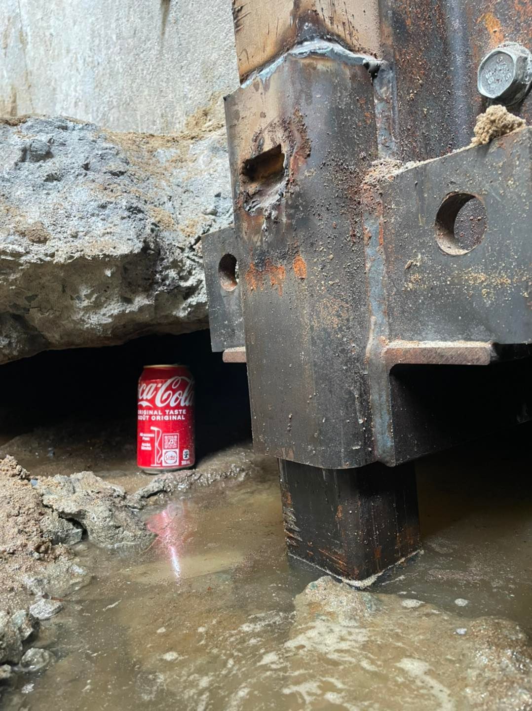 Une canette de Coca-Cola repose dans un trou boueux creusé sous une lourde poutre de soutien en acier rouillé.