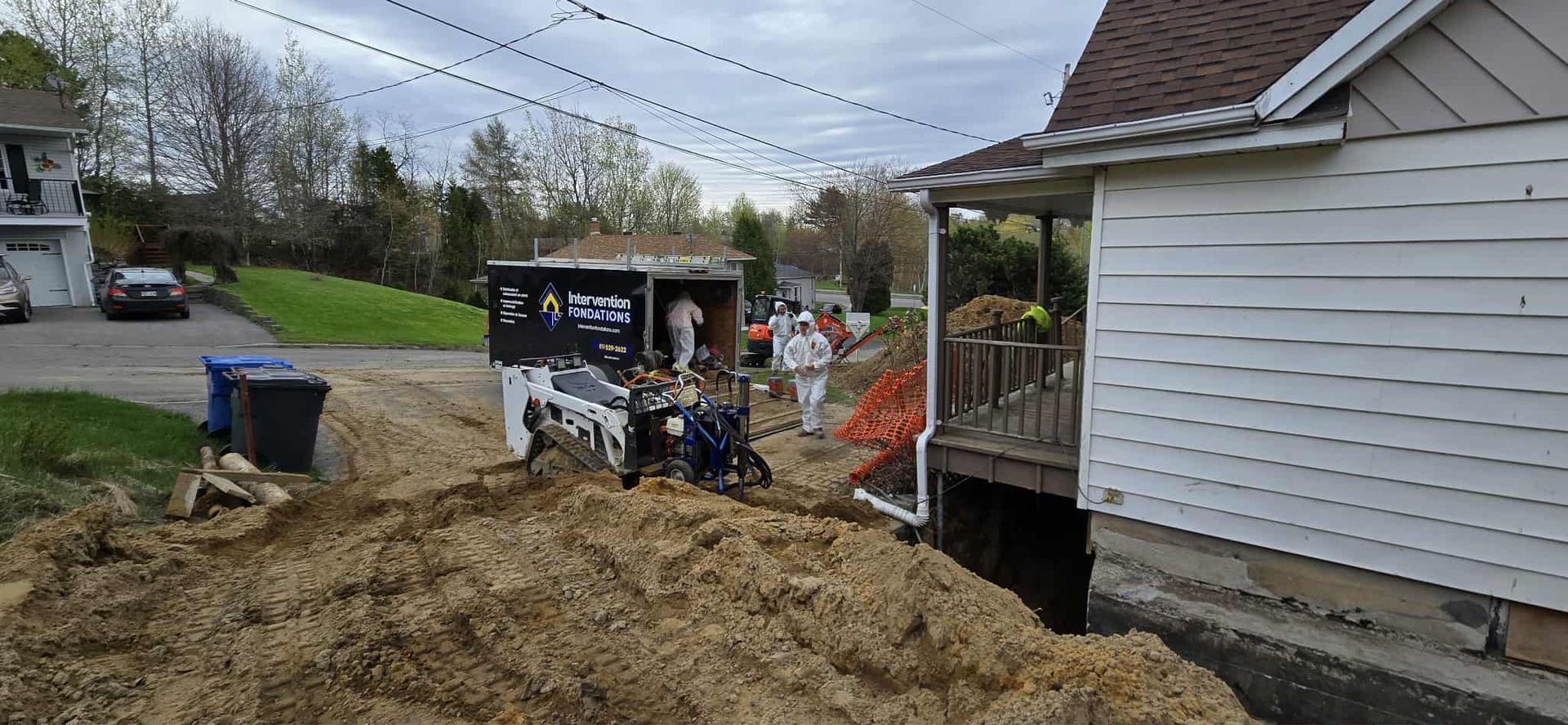 Une chargeuse compacte creuse une tranchée à côté d'une maison tandis que des ouvriers en combinaisons de protection blanches se tiennent à proximité.