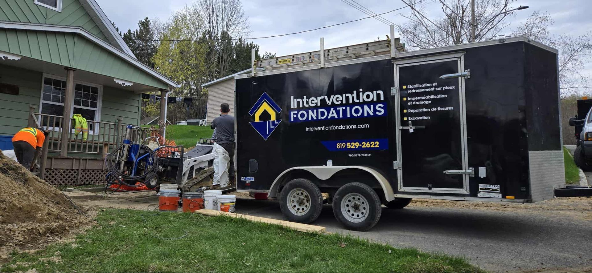 Des ouvriers du bâtiment travaillent sur les fondations d'une maison à côté d'une remorque de chantier noire de la société « Intervention Fondations ».