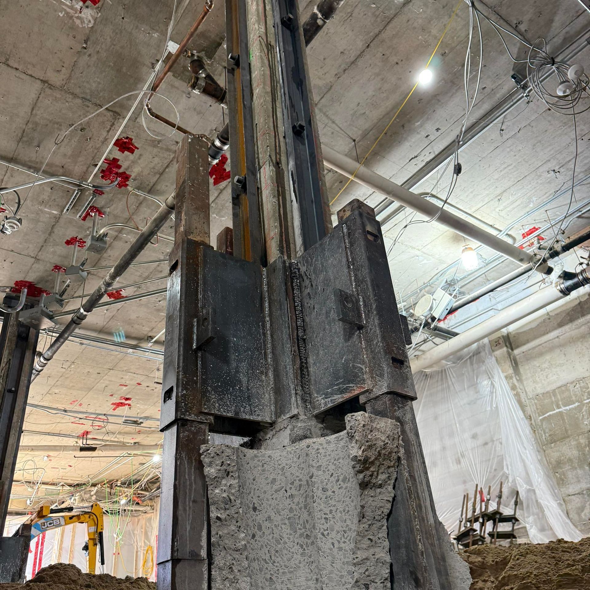 Une lourde colonne de soutien en acier, avec des plaques métalliques et une fondation en béton apparente, sur un chantier de construction intérieur.