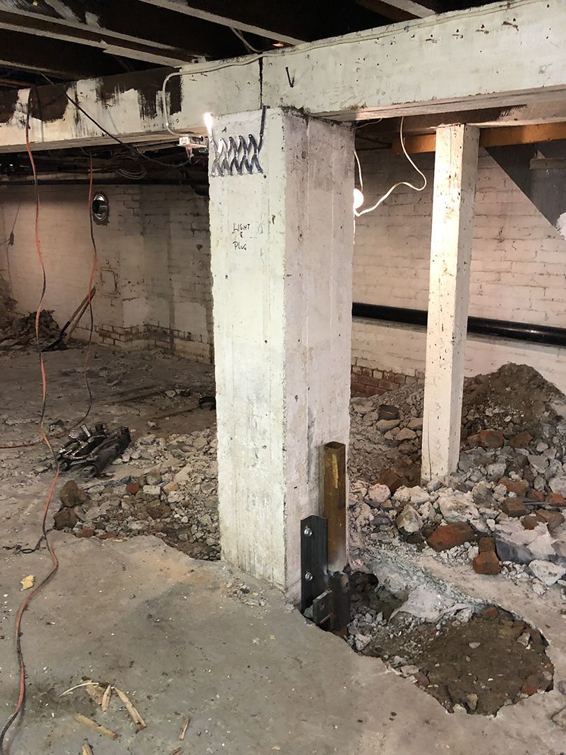 L'intérieur d'un sous-sol montre deux colonnes verticales en béton blanc soutenant une poutre, avec de la terre excavée autour des bases.