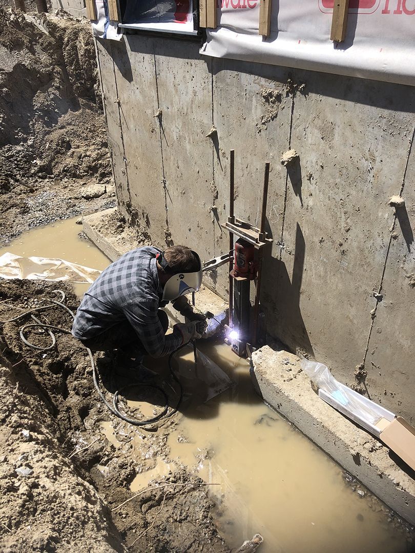 Un ouvrier portant un masque de soudeur utilise un chalumeau pour souder un support de pilier métallique à une fondation en béton dans une tranchée boueuse.