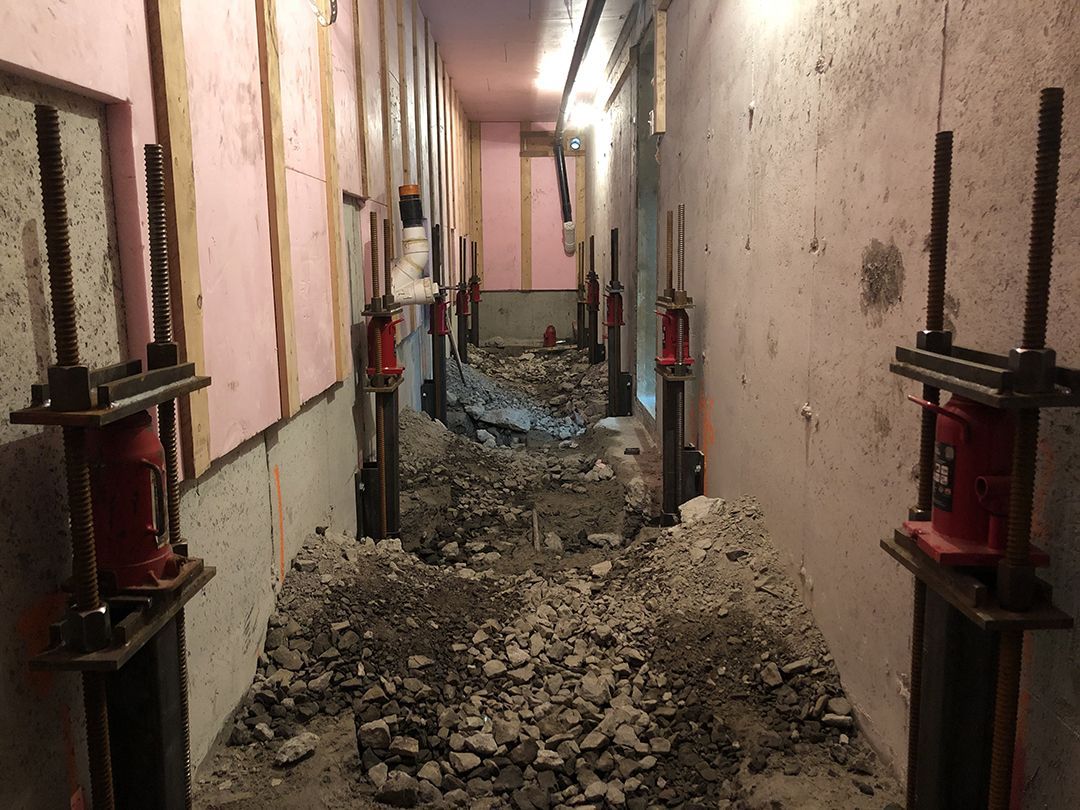 Un sous-sol étroit en construction, avec plusieurs vérins hydrauliques rouges soutenant une poutre le long de murs en béton.