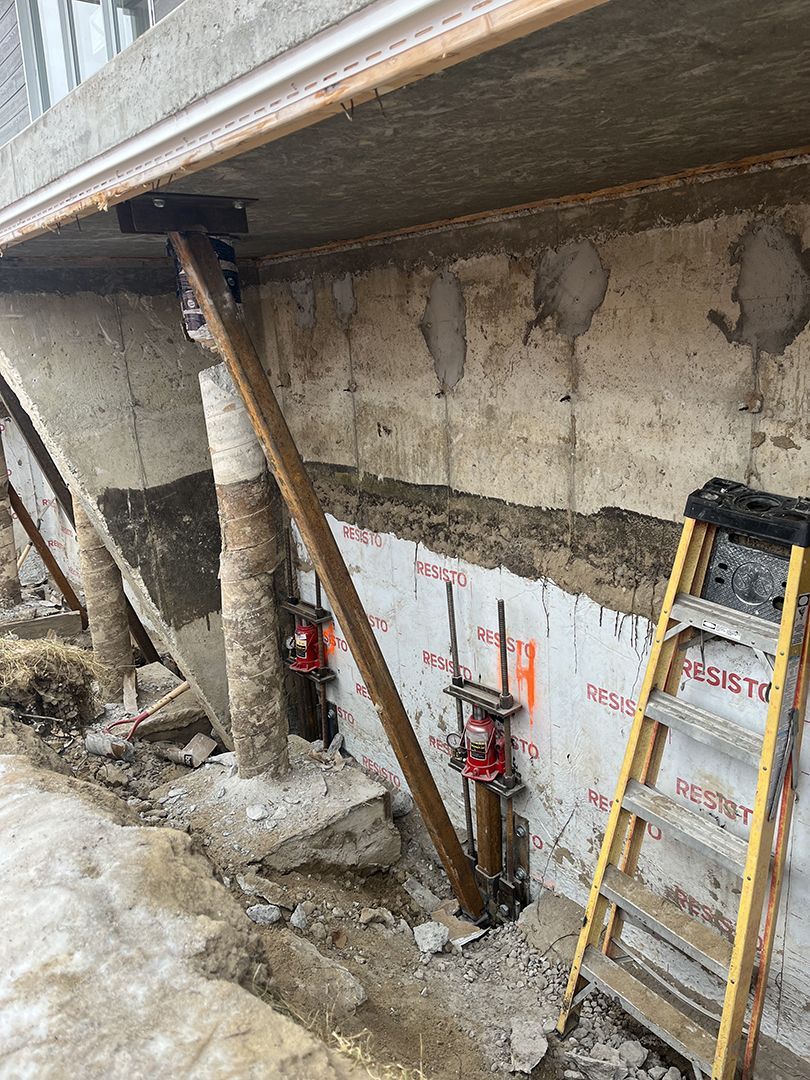 Chantier de construction montrant la réparation de fondations en béton avec des supports verticaux en acier, des contreventements diagonaux et une échelle jaune.