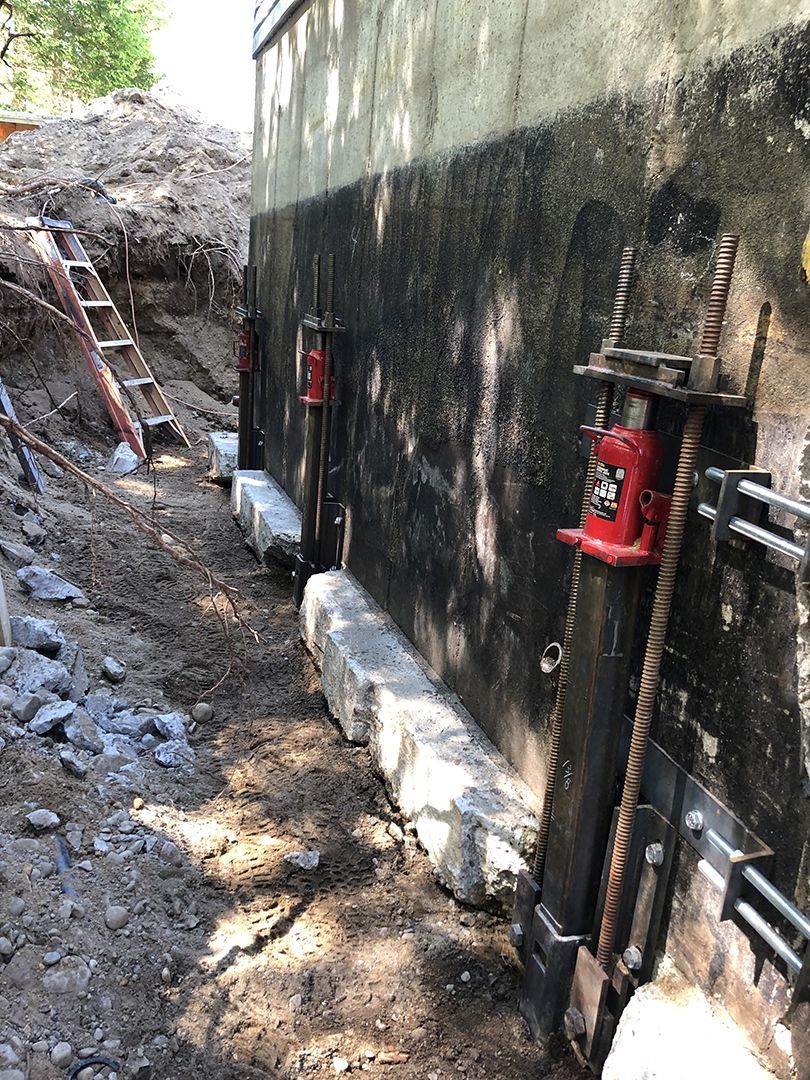 Des vérins hydrauliques robustes sont installés contre le mur de fondation d'une maison, dans une tranchée profonde creusée dans le sol.