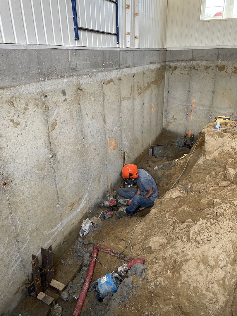 Une personne portant un casque de chantier, agenouillée dans une tranchée de terre à côté d'un mur de fondation en béton, travaille sur des tuyaux et des câbles apparents.