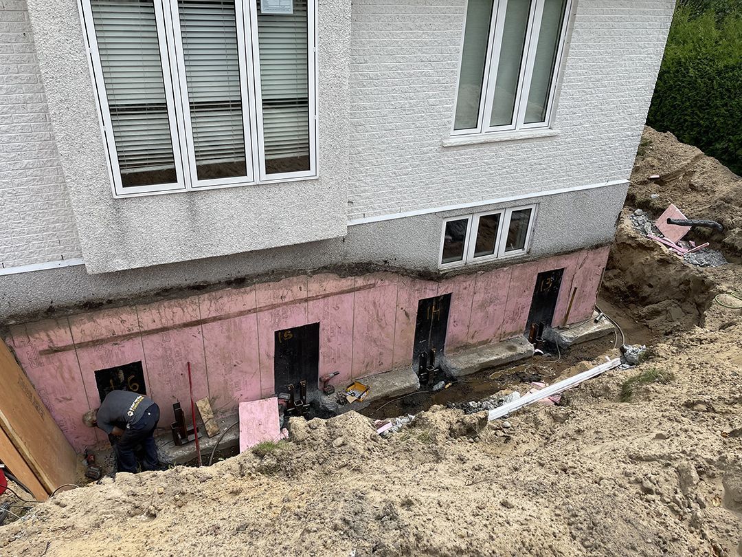Un ouvrier répare, dans une tranchée, les fondations d'une maison contre un mur extérieur isolé en rose, sous de grandes fenêtres au niveau du sol.