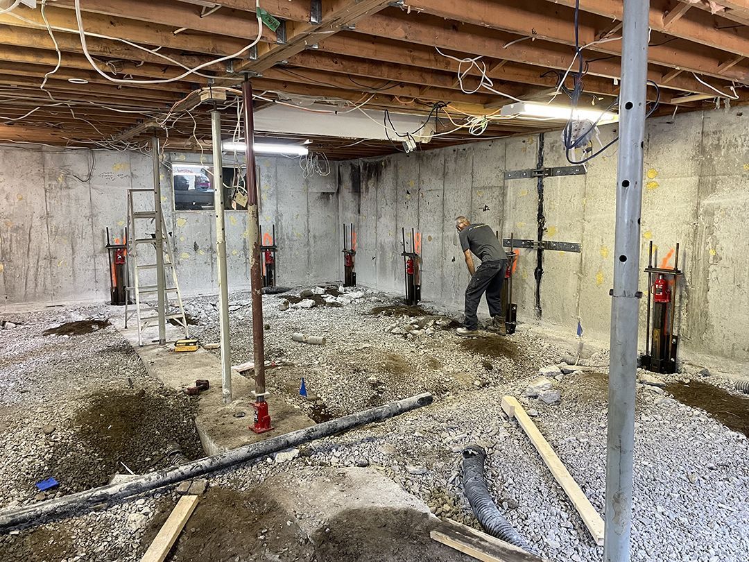 Un ouvrier du bâtiment installe des étais métalliques dans un sous-sol dont le sol est recouvert de gravier et les solives apparentes.