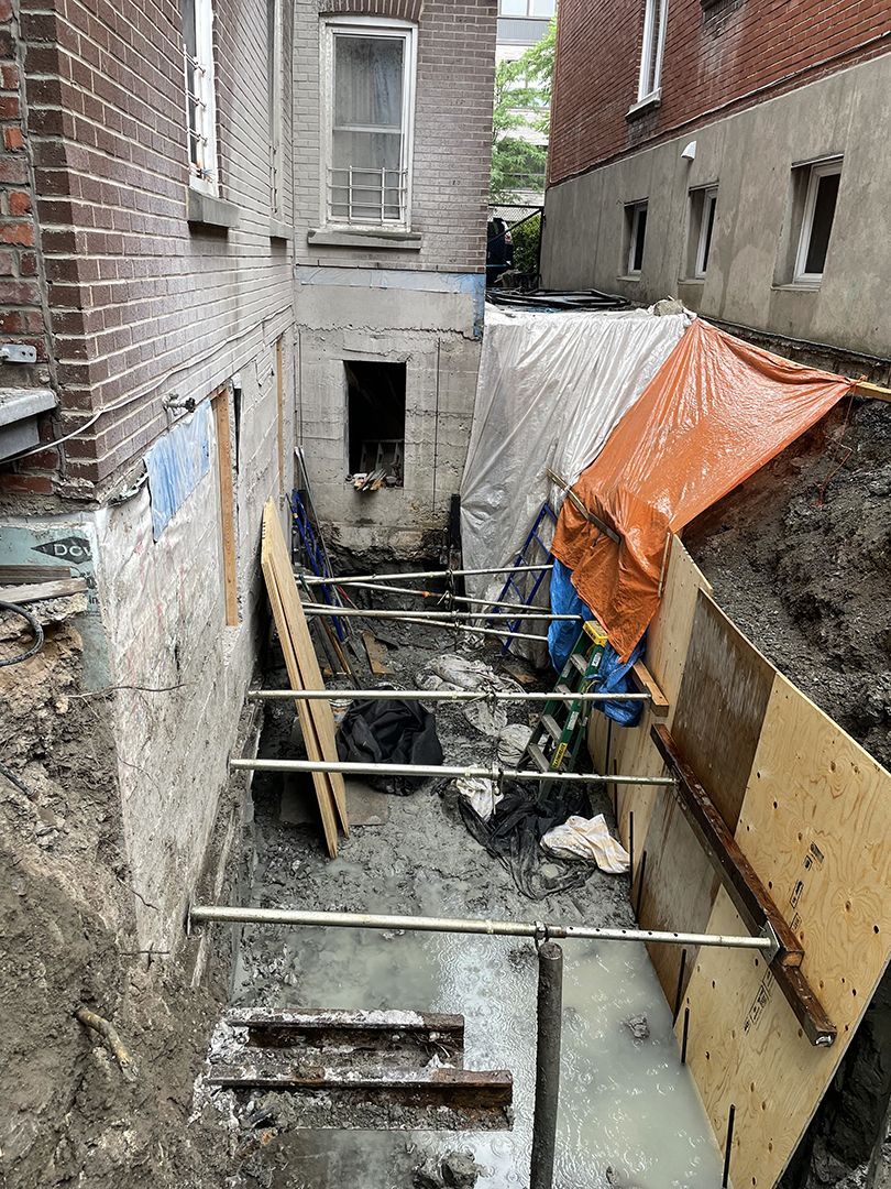 Une étroite tranchée de construction entre deux bâtiments en briques, étayée par des poteaux en acier et du contreplaqué, au sol boueux et jonché de débris.