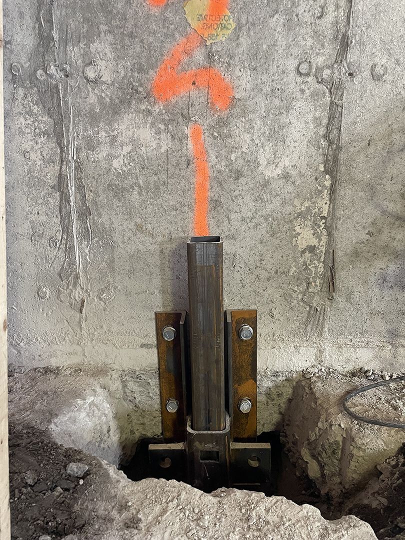 Un support de pilier en acier est boulonné à un mur de fondation en béton, avec des marquages ​​de construction orange au-dessus.