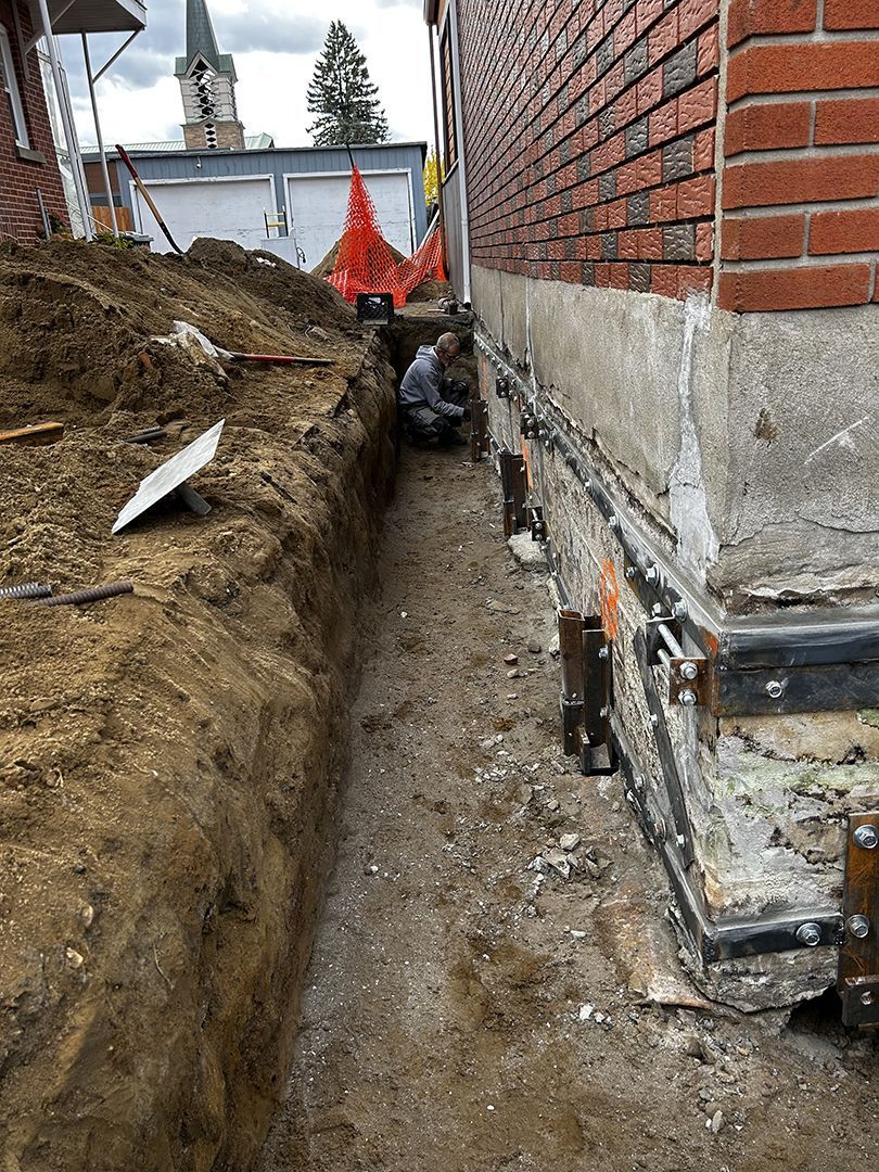 Un ouvrier répare les fondations d'une maison en briques, dans une tranchée de terre creusée à côté de celle-ci.